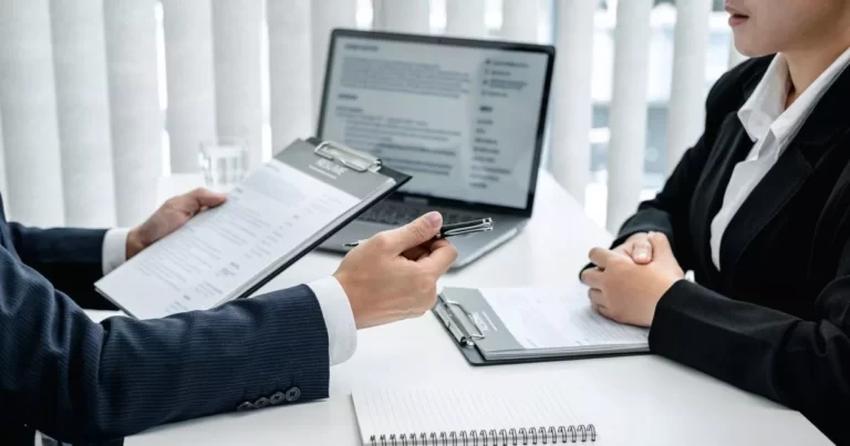 Man at desk holding clipboard, speaking to woman sitting on other side of desk, representing frequently asked questions