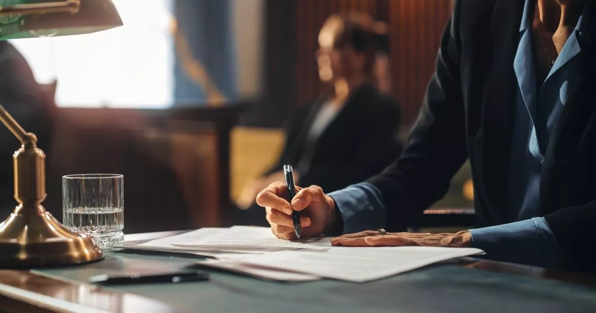 Woman in suit sitting at table, holding a pen with a glass of water and a lamp. Image represents investigation services for lawyers