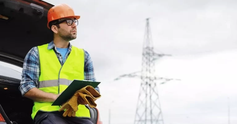 Worker in orange hard hat and yellow high-vis vest, leaning against car and holding a clipboard, with electricity transmission tower in background. Image represents workplace policies and systems for safety issues